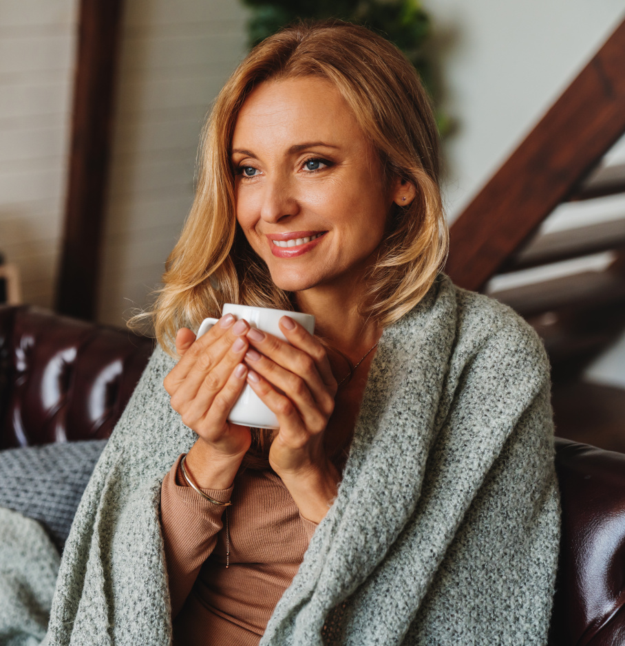 Close up of dreaming middle aged woman sitting in living room with cup of coffee or tea enjoying under blanket